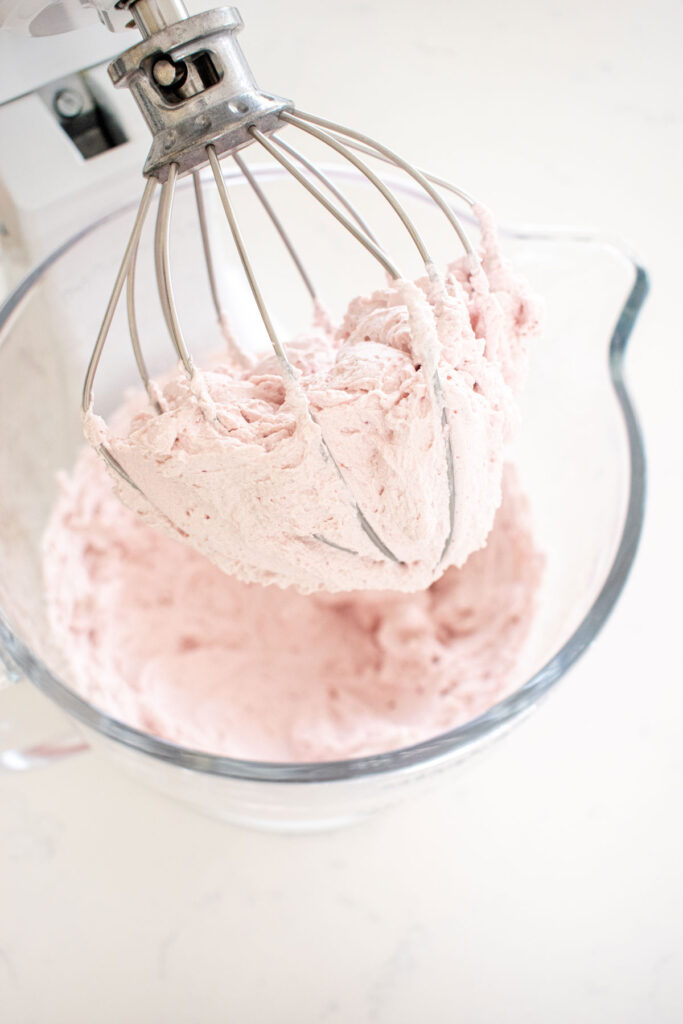 whipped heavy cream and raspberry powder whipped into stiff peaks in a glass stand mixing bowl on a white marble counter.