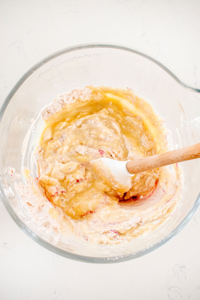 sweetened condensed milk being added into raspberry whipped cream in a glass mixing bowl on a white marble counter.