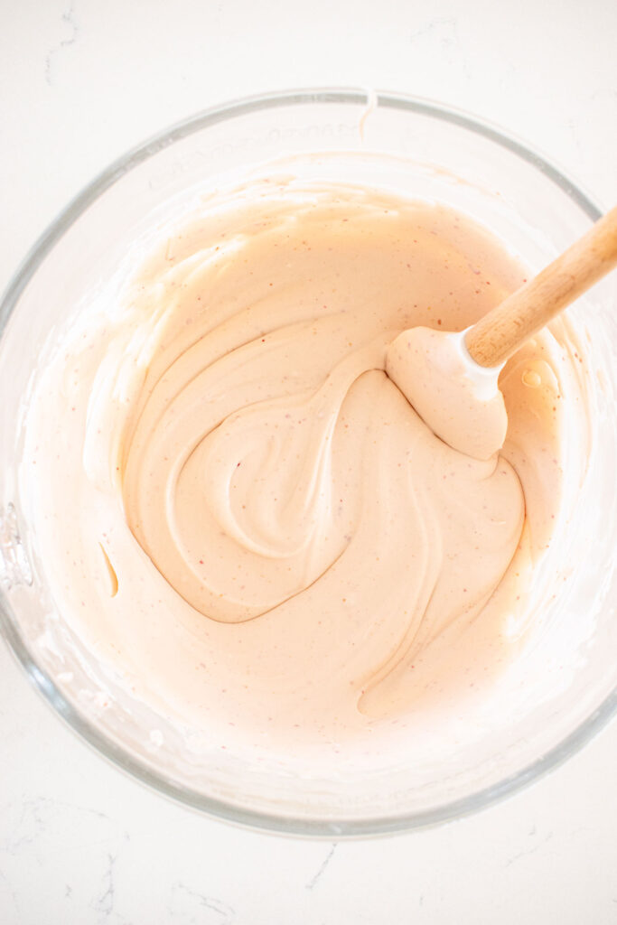 raspberry no churn ice cream in a glass mixing bowl with a spatula on a white marble counter.