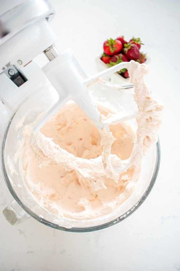 fresh strawberry buttercream frosting in a glass mixing bowl on a white marble countertop.