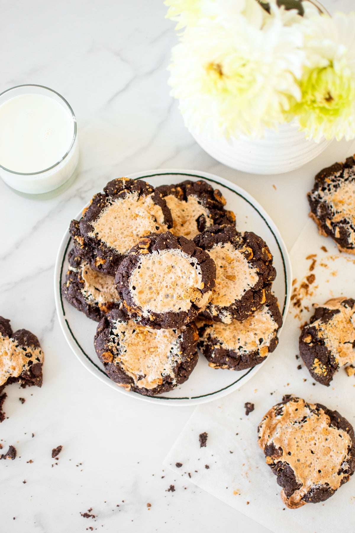 bakery style chocolate smores cookies with fluff on a plate on a white marble countertop with a glass of milk and flowers.