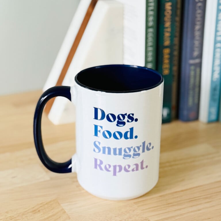 dogs food snuggle repeat mug on a wood counter with books behind it.