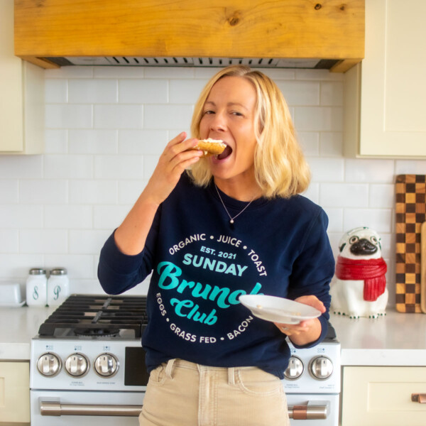 woman wearing brunch club sweatshirt standing in kitchen eating a bagel.