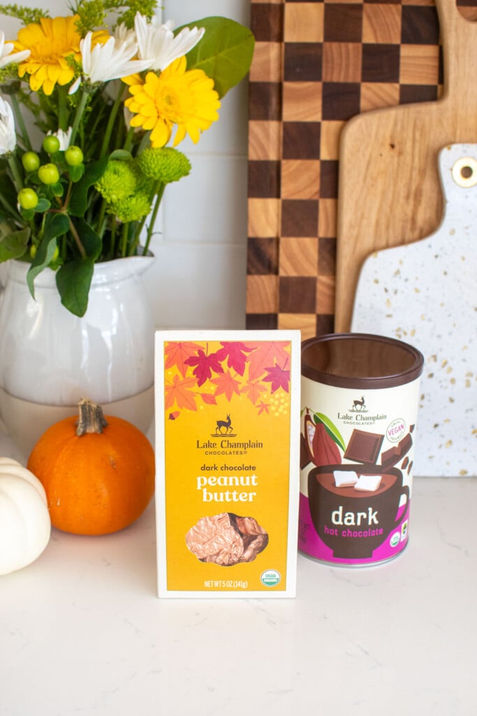 lake champlain dark hot chocolate mix and peanut butter chocolate leaves on a white marble counter with fresh flowers and pumpkins.