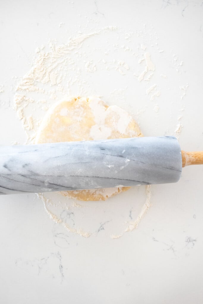 pie crust being rolled out with a marble rolling pin on a white marble counter.