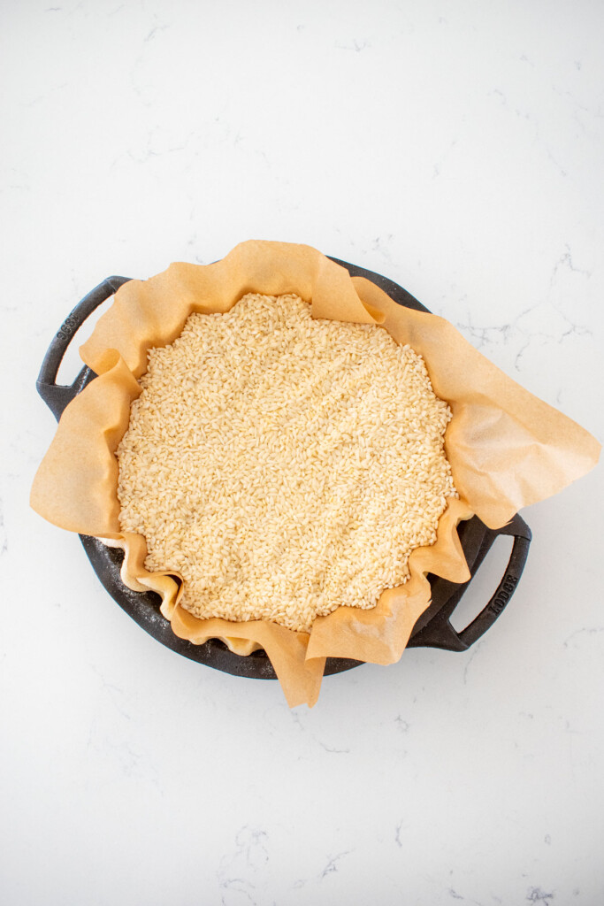 fluted pie crust with parchment paper and pie weights in a cast iron pie pan on a white marble counter.