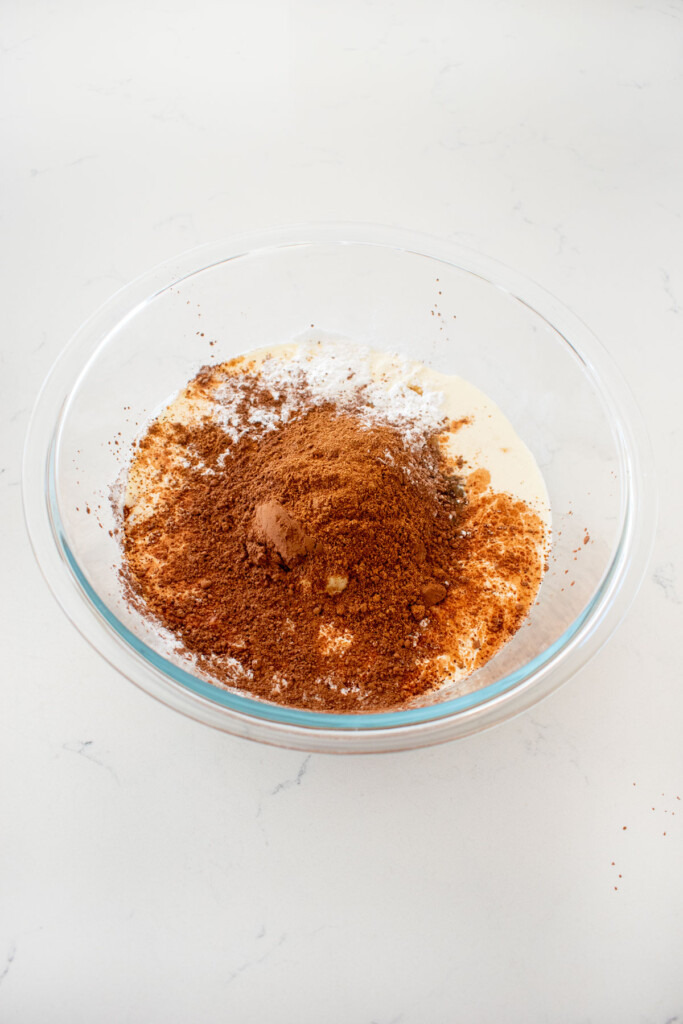 ingredients for chocolate pumpkin pie filling in a glass mixing bowl on a white marble counter.