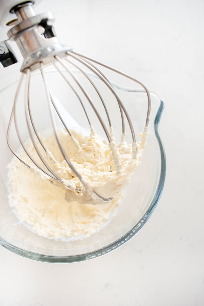 homemade whipped cream in a glass mixing bowl with a whisk on a white marble counter.
