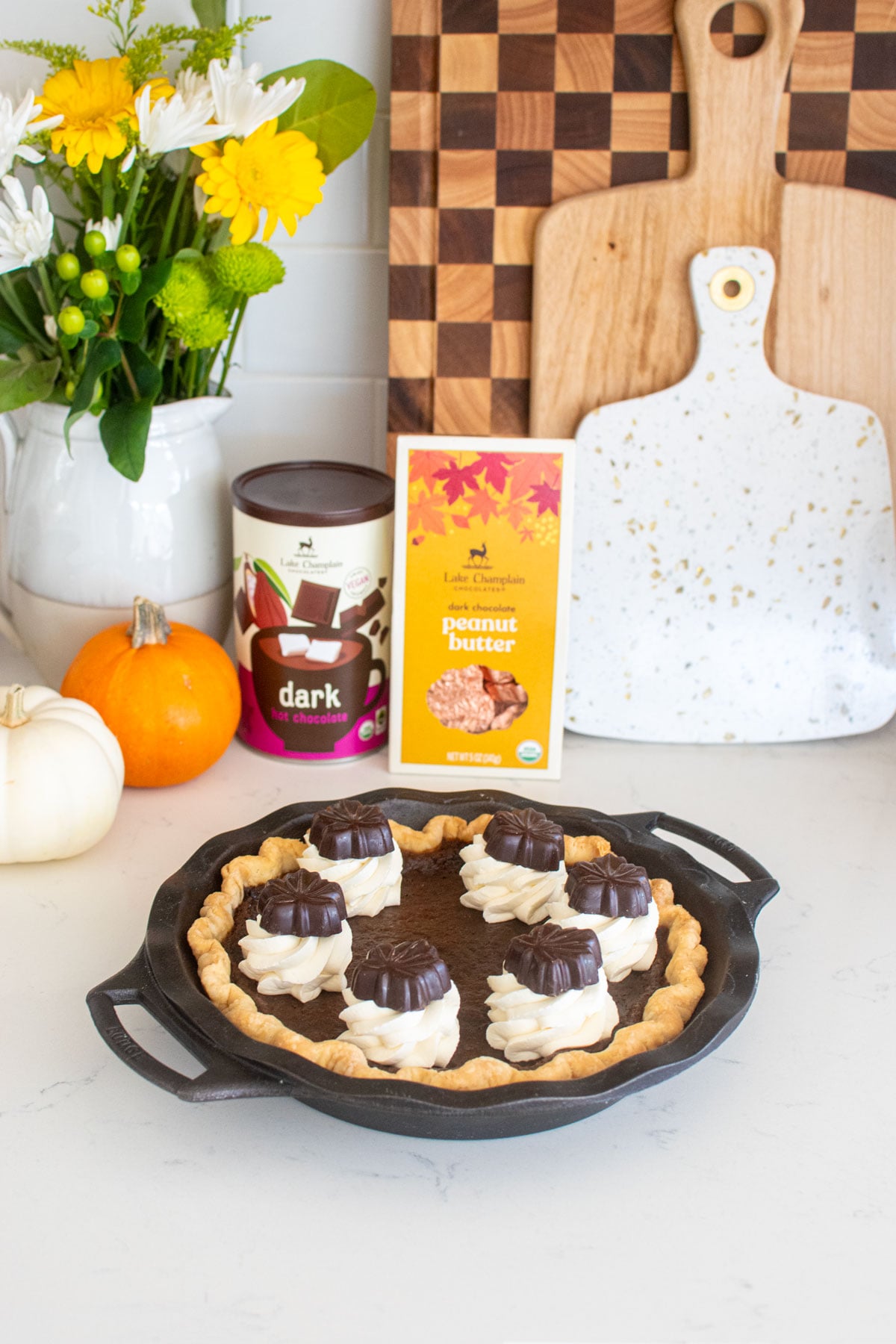 chocolate pumpkin pie without eggs with whipped cream dollops and chocolate leaves on top in a cast iron pie pan on a white marble counter with pumpkins and fresh flowers.
