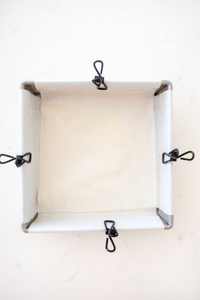 8x8-inch baking pan lined with parchment paper and rubbed with butter being held up by binder clips on a marble counter.