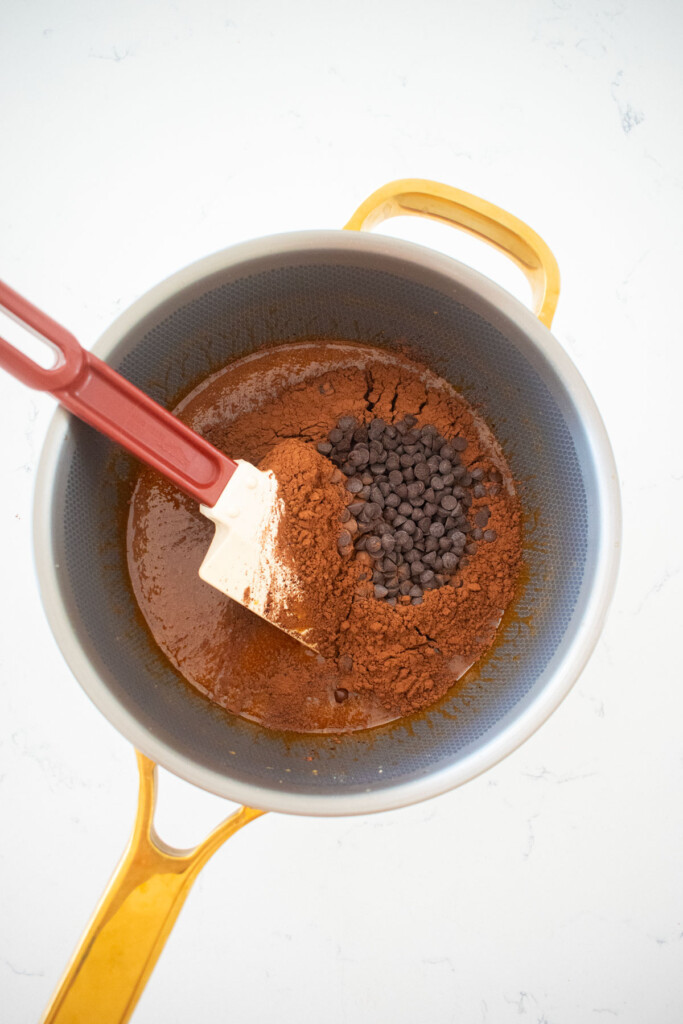 dark chocolate and cocoa powder being added to caramel in a stainless steel pot on a marble counter.