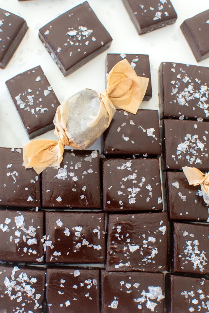 homemade chewy chocolate caramel candy with flaky sea salt cut into squares on a white marble counter.