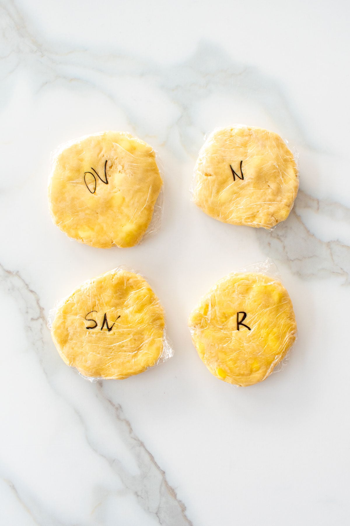 four small pie doughs wrapped in plastic wrap on a white marble counter with letters on them indicating which butter was used to make them.