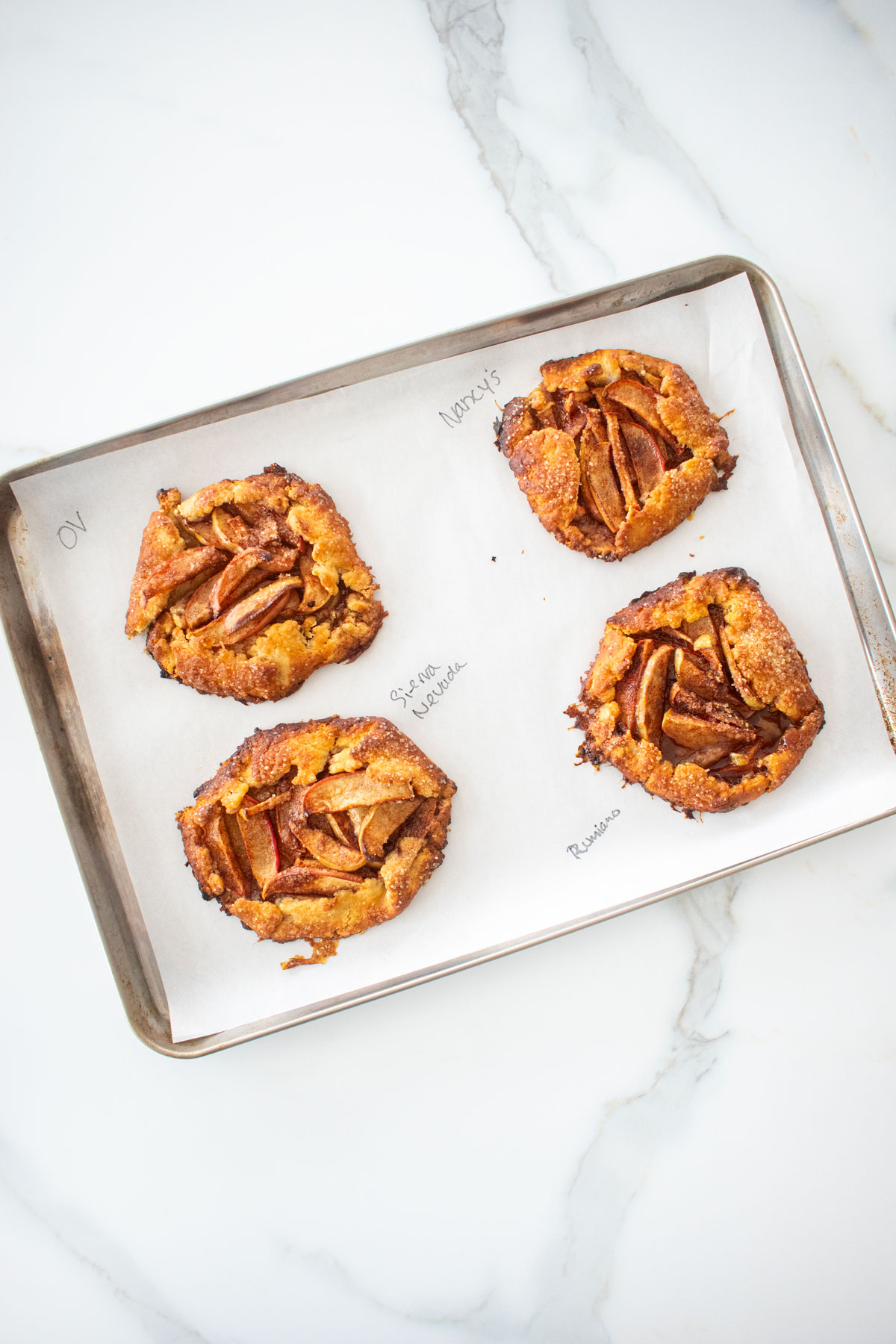 mini apple galettes on a baking sheet with the butter name used written on the parchment paper on a white marble counter.
