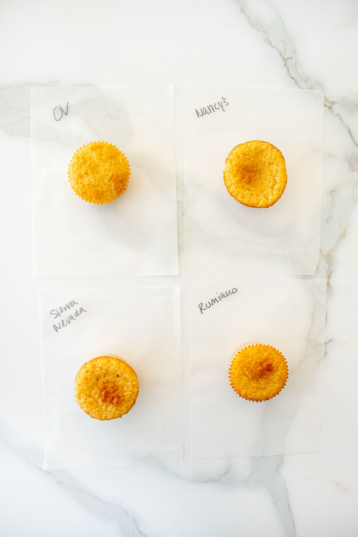 four vanilla cupcakes on a piece of white parchment paper made with different butters with the name written in black marker on a marble counter.