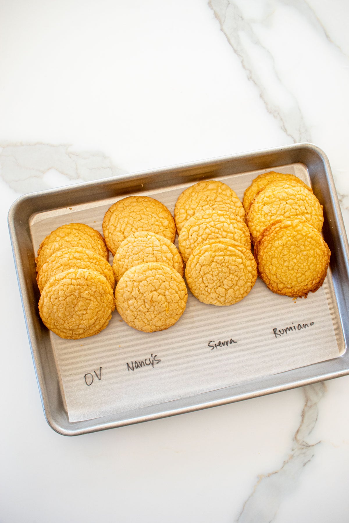 four different vanilla shortbread cookies baked on a baking sheet with labels for the different butter used to test them on a white marble counter.
