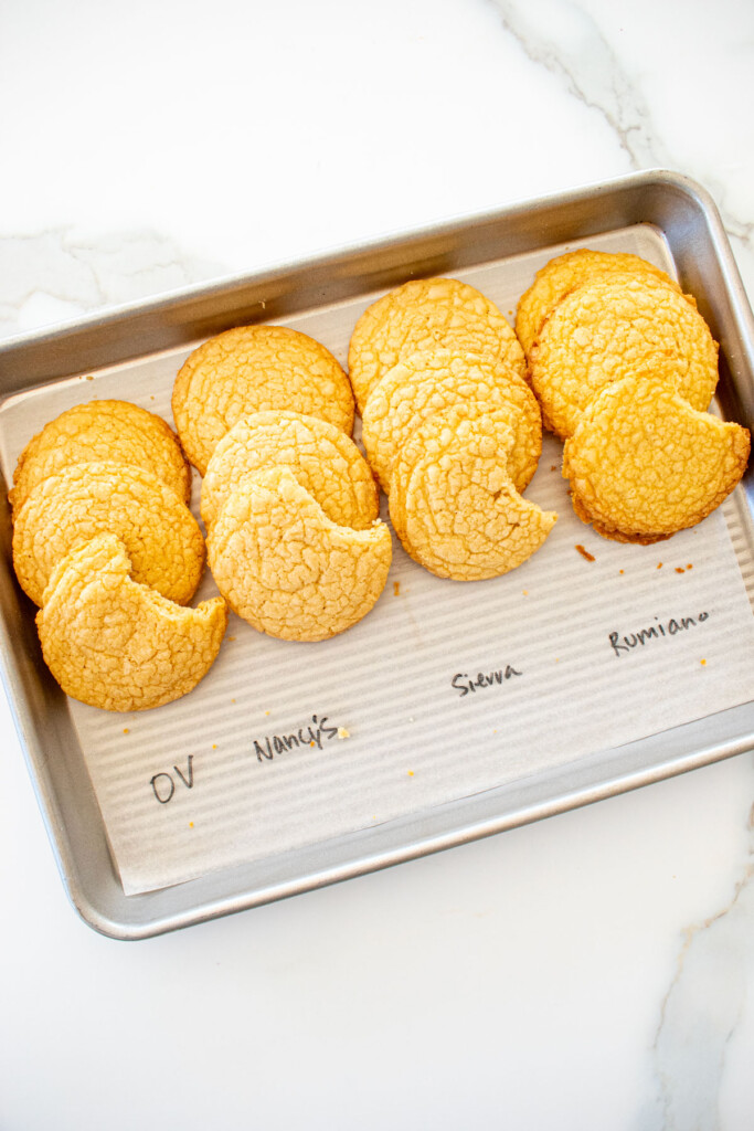 four different vanilla shortbread cookies baked on a baking sheet with labels for the different butter used to test them on a white marble counter.