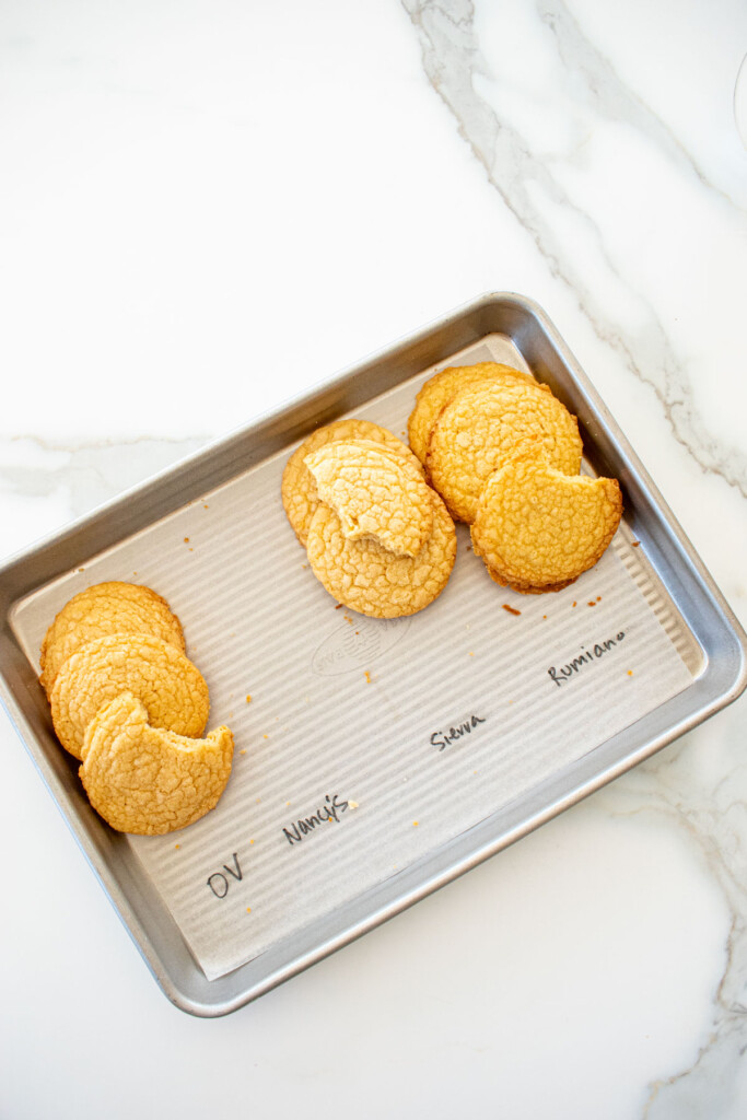four different vanilla shortbread cookies baked on a baking sheet with labels for the different butter used to test them on a white marble counter.