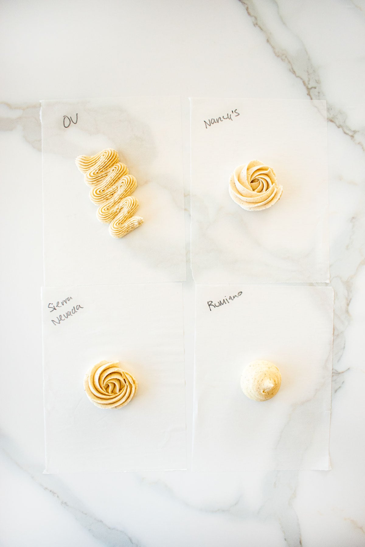 four different vanilla frostings piped onto parchment paper with the butter name that was used written in black ink on a white marble counter.