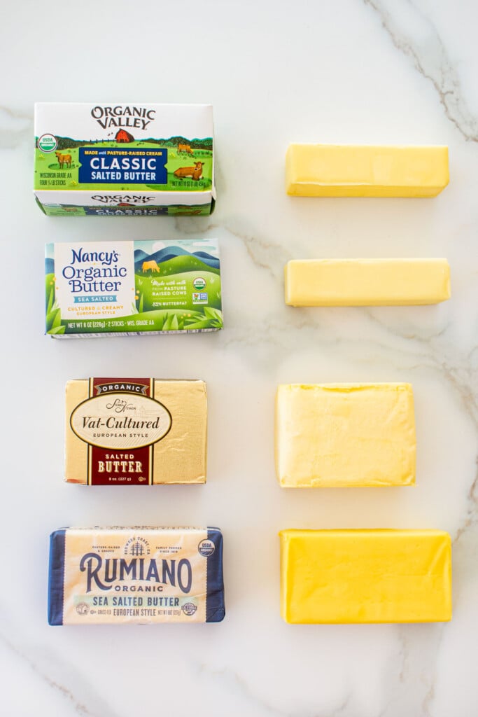 four top butters side by side on a marble countertop showing the brand box and then the butter opened next to it to see color and texture.