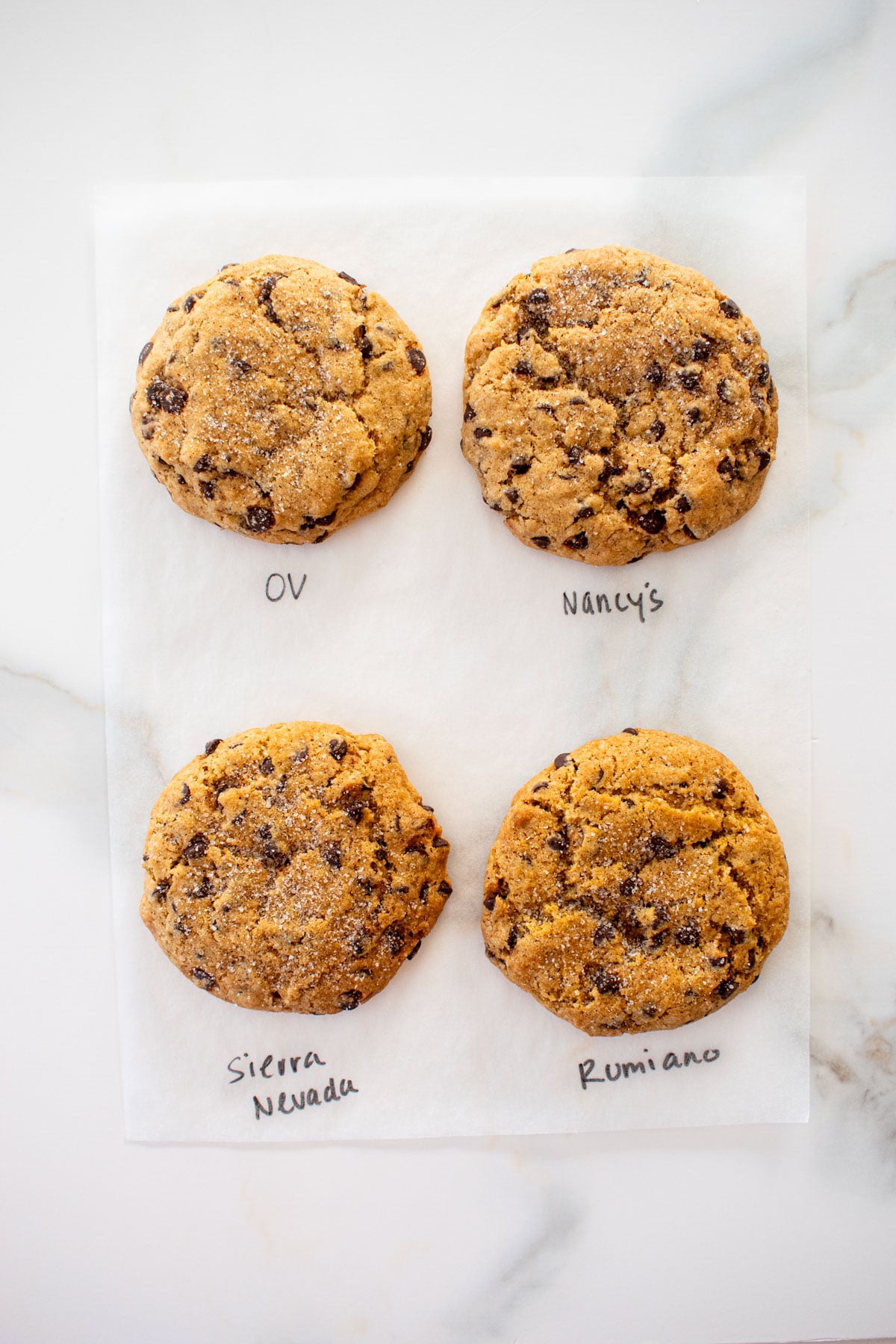 four chocolate chip cookies made with four different butters on a white marble counter with the butter name written next to each cookie.