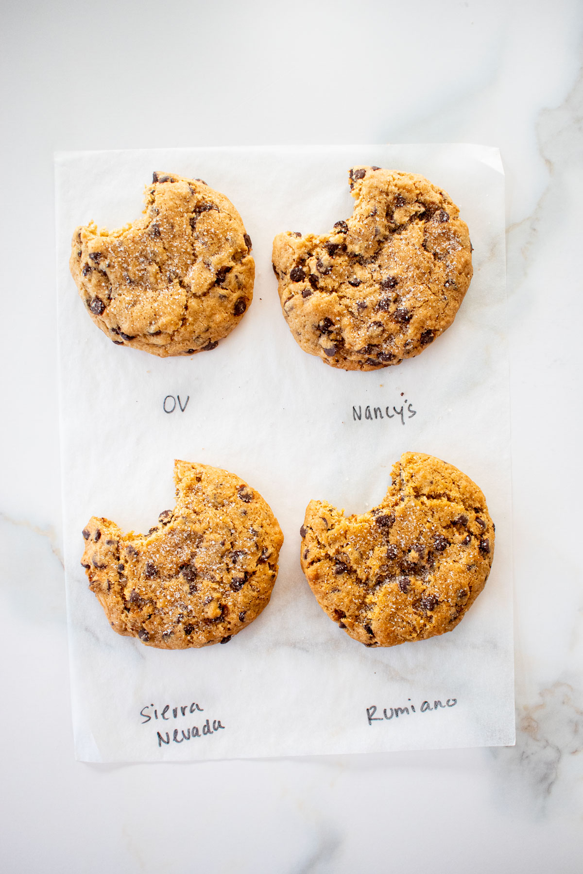 four chocolate chip cookies made with four different butters with a bite taken out of each on a white marble counter with the butter name written next to each cookie.