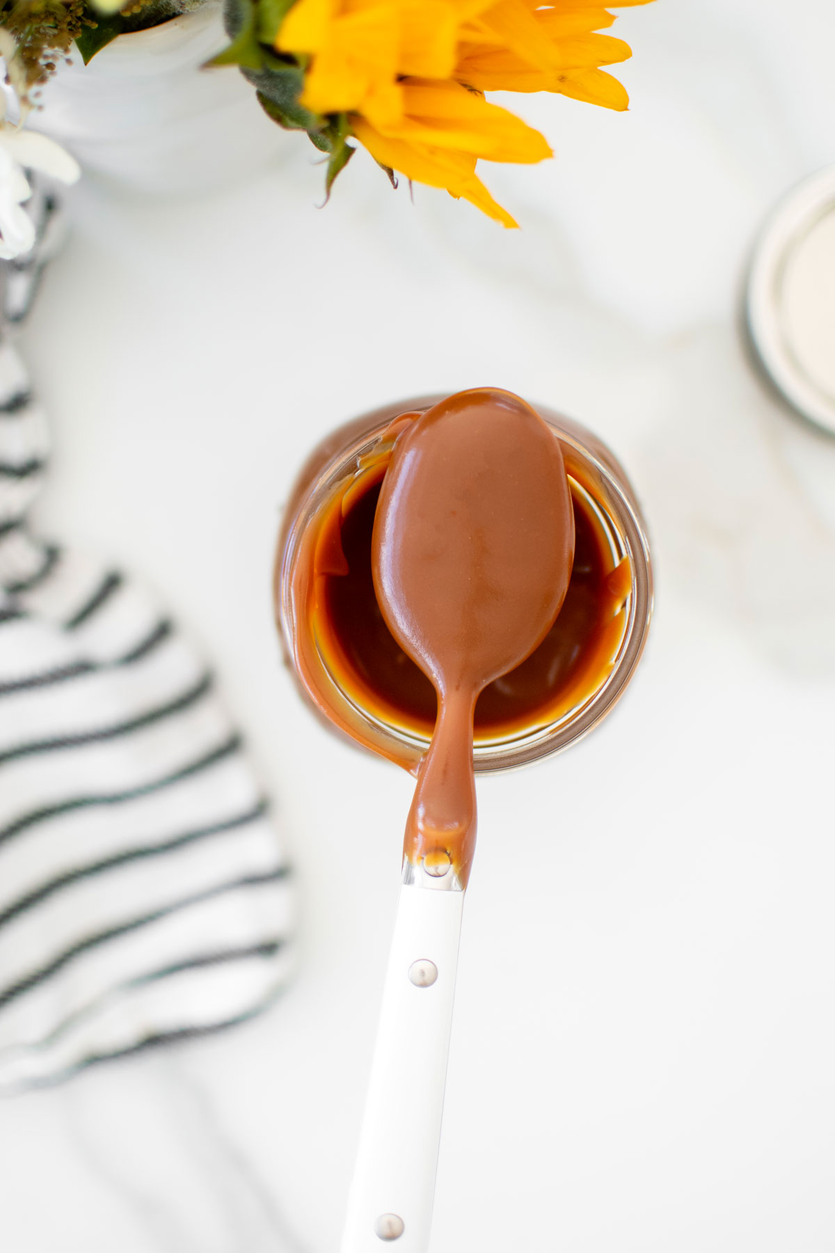 homemade true caramel sauce on a spoon in a mason jar on a marble counter with flowers.