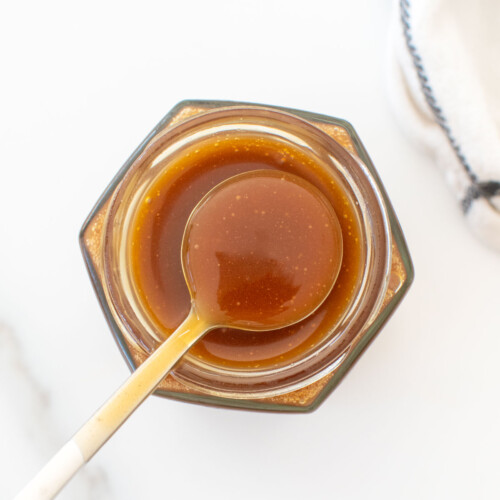 homemade caramel sauce with bone broth on a spoon over a glass jar on a white marble counter.