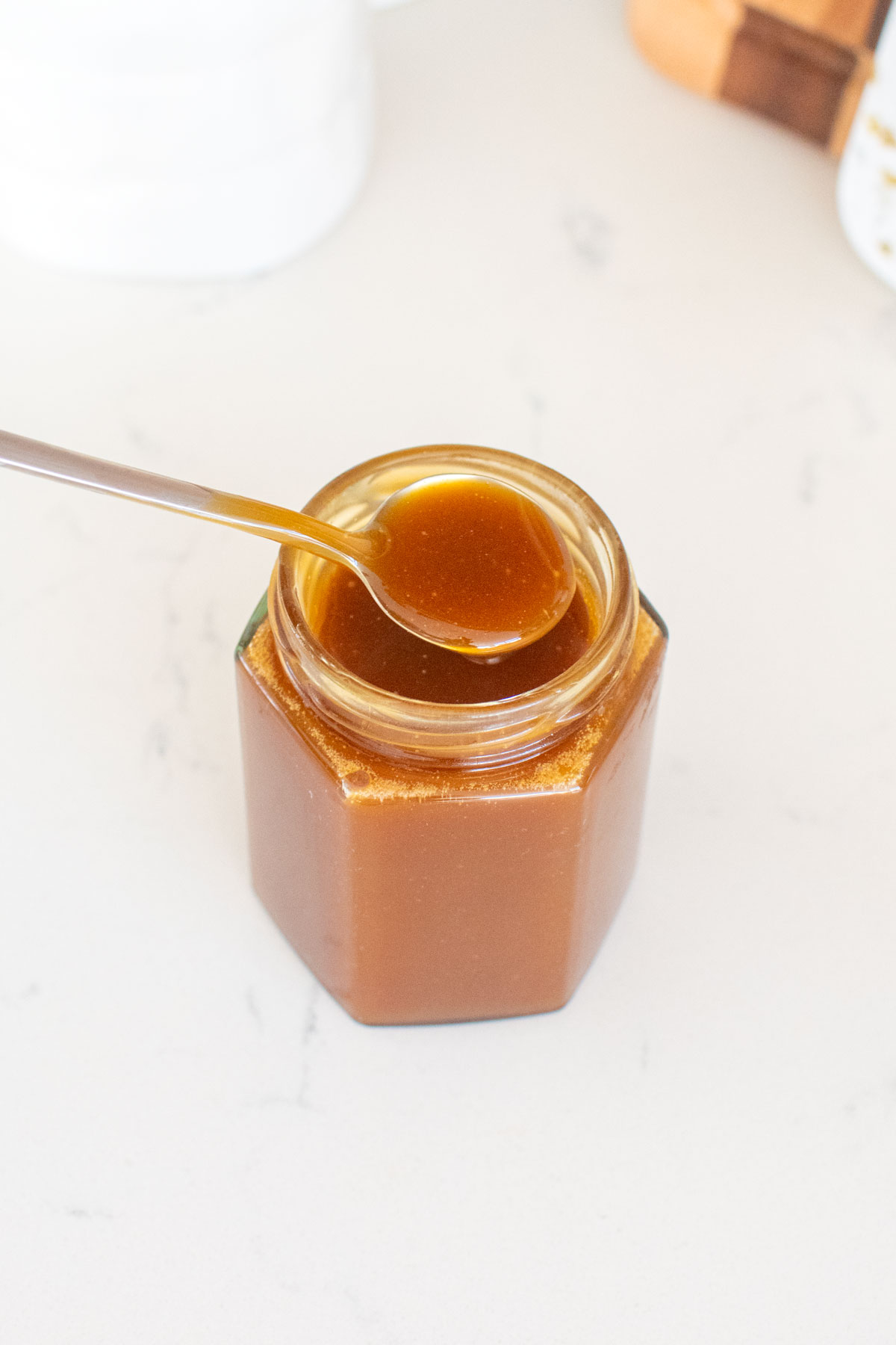 homemade caramel sauce with bone broth on a spoon over a glass jar on a white marble counter.