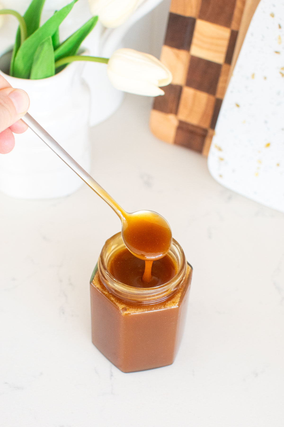 homemade caramel sauce with bone broth on a spoon over a glass jar on a white marble counter.