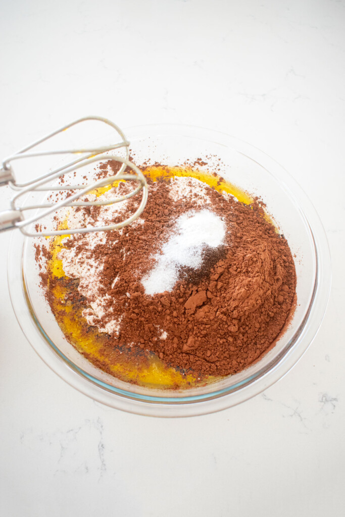 dry ingredients added into eggless chocolate cake batter in a glass mixing bowl on a white marble counter.