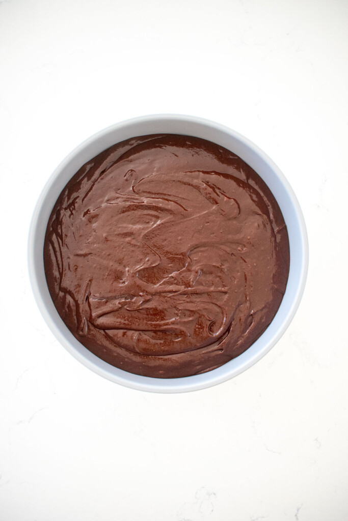 eggless chocolate cake batter in a round cake pan on a white marble counter.