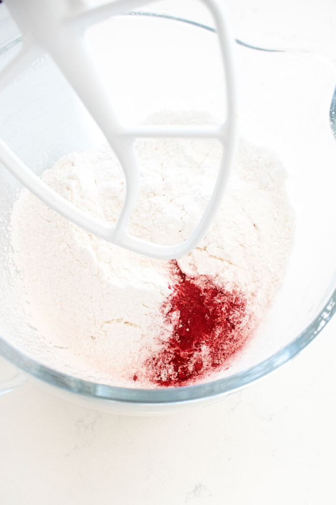 ingredients for pink vanilla frosting in a glass mixing bowl on a white marble counter.
