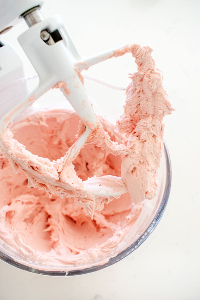 light pink frosting in a glass mixing bowl on a white marble counter.