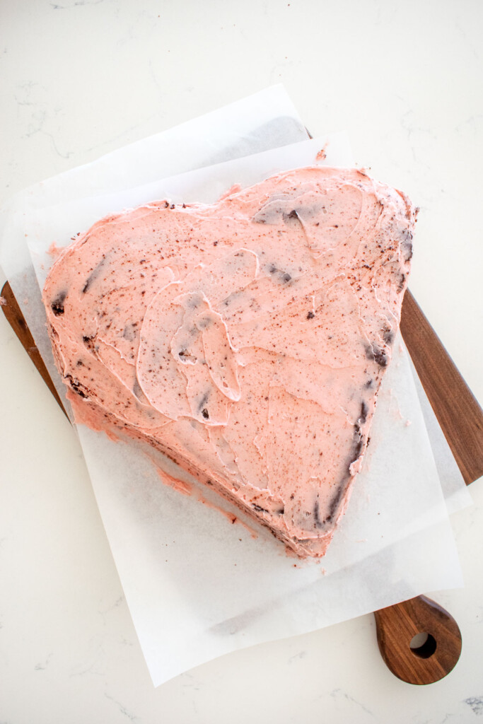 eggless chocolate heart cake crumb coated in pink frosting on a wood board on a marble counter.