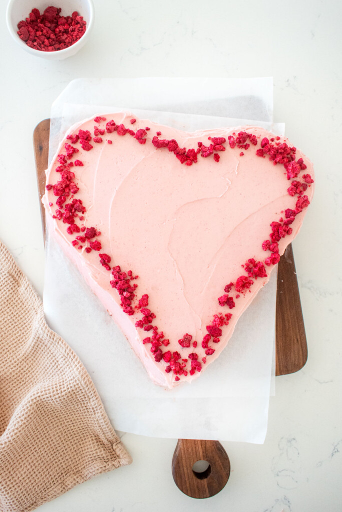 eggless chocolate heart cake with pink frosting and freeze dried raspberries on a white marble counter.