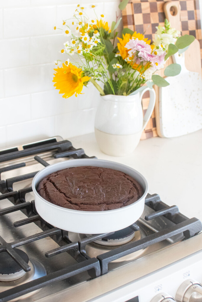 baked eggless chocolate cake in a round cake pan cooling on top of an oven.