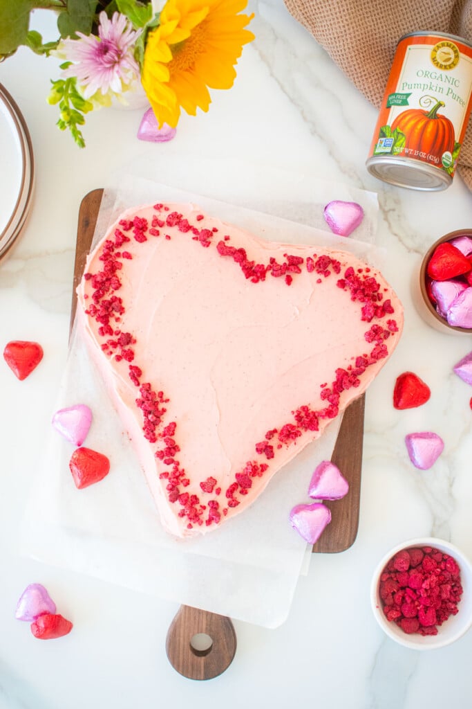 chocolate heart cake frosted with pink frosting and decorated with freeze dried raspberries on a marble counter with flowers and chocolate hearts.
