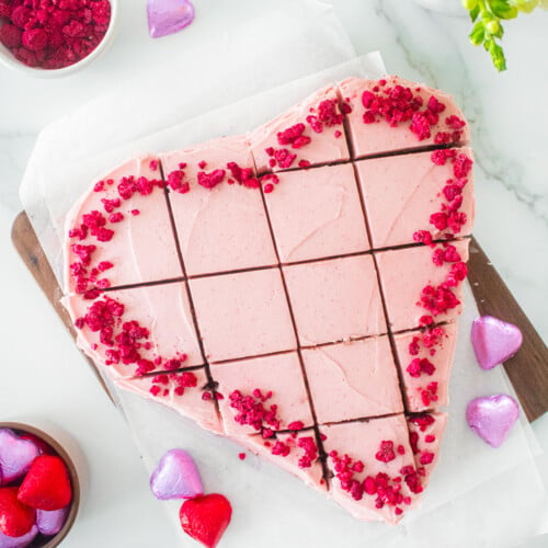 eggless chocolate heart cake with pink frosting and freeze dried raspberries cut into squares on a white marble counter.
