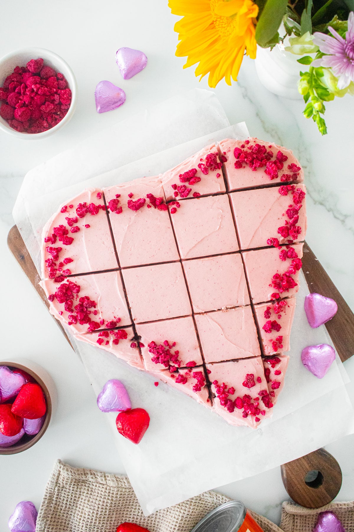 eggless chocolate heart cake with pink frosting and freeze dried raspberries cut into squares on a white marble counter.