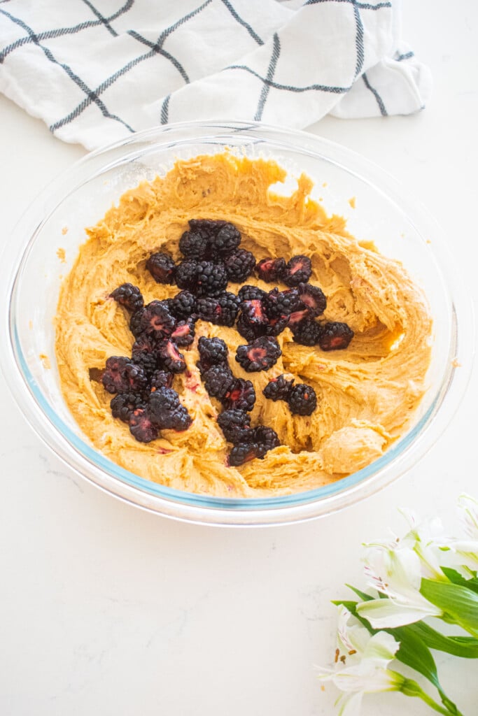 sweet potato muffin batter in a glass mixing bowl with fresh blackberries on a white marble counter.