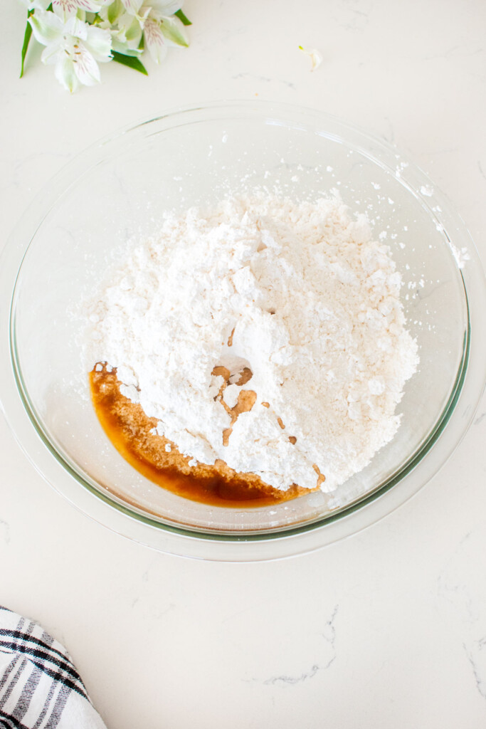large glass mixing bowl with peanut butter, bone broth, coconut flour, powdered sugar and vanilla on a white marble counter.