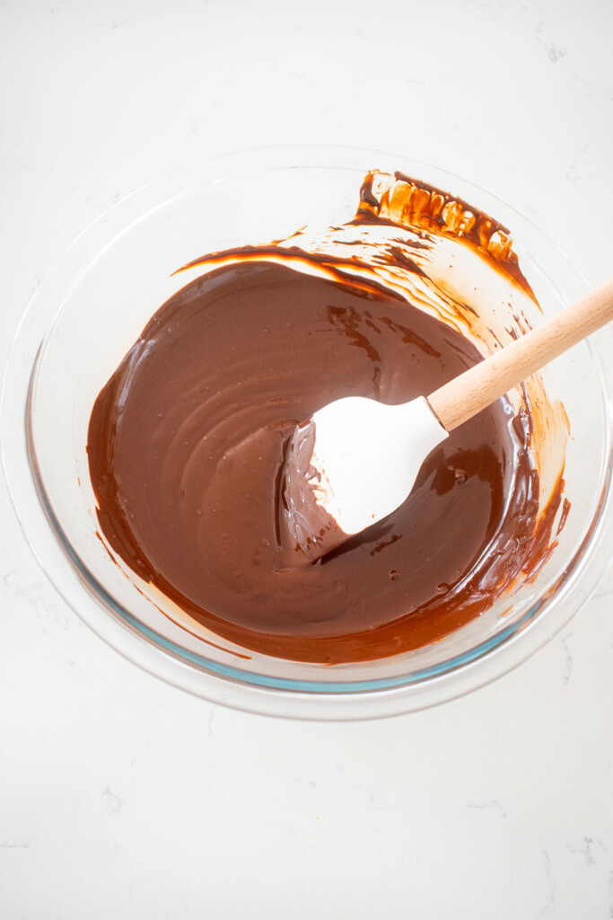glass mixing bowl with melted chocolate and a spatula on a white marble counter.