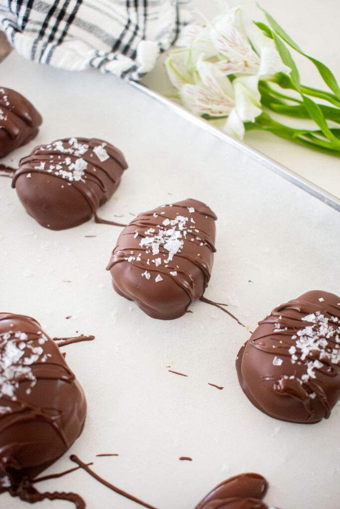 chocolate peanut butter eggs with chocolate drizzle and flaky sea salt on top on a baking sheet on a white marble counter.