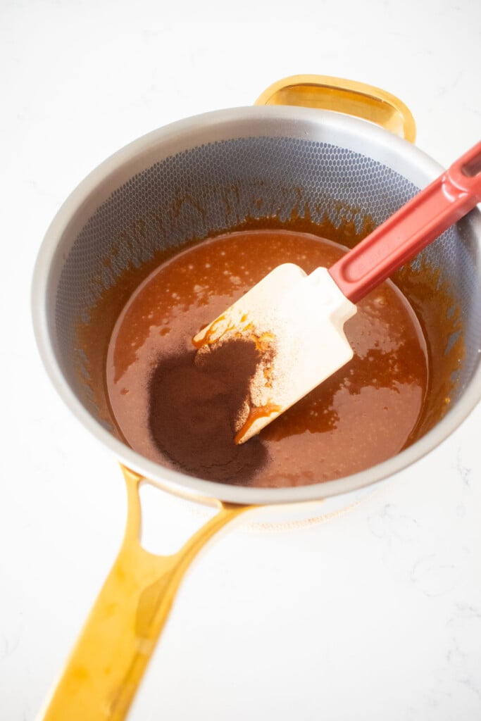 espresso powder being added to caramel in a saucepan on a white marble counter.