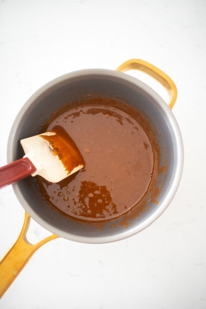 homemade chewy coffee caramel being stirred in a saucepan with a spatula on a white marble counter.
