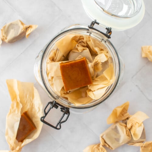 homemade chewy coffee caramel candies wrapped in wax paper in a glass canister on a white tile table.