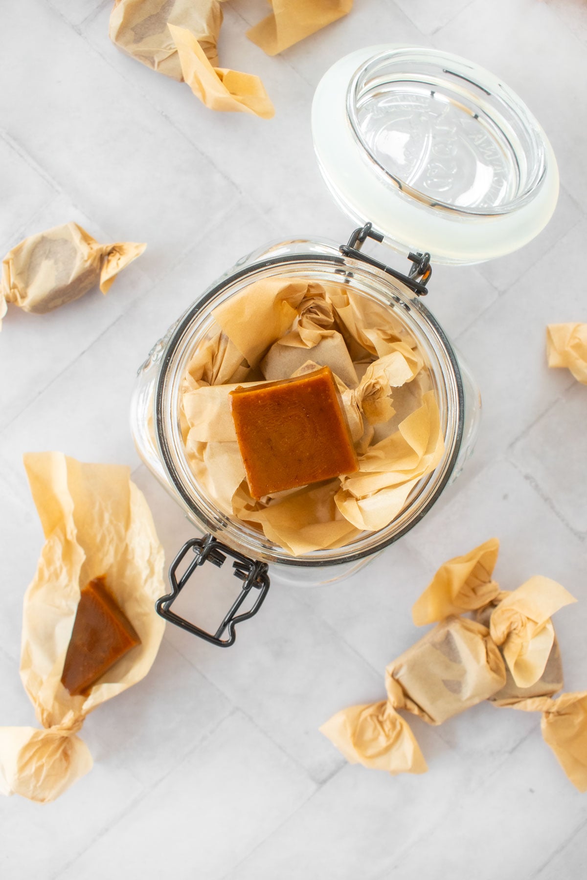 homemade chewy coffee caramel candies wrapped in wax paper in a glass canister on a white tile table.