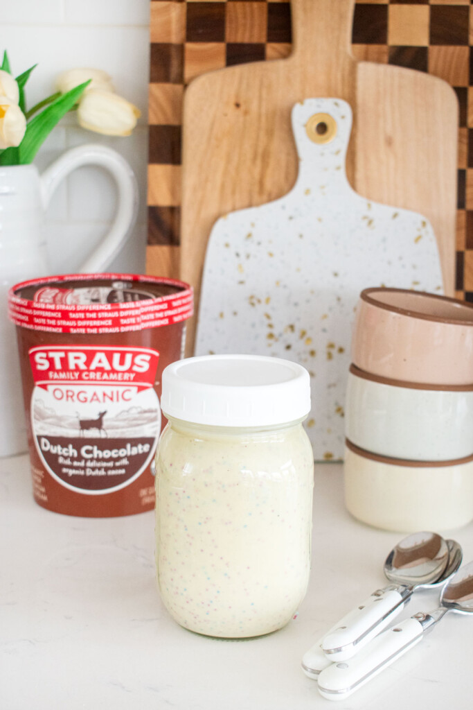 mason jar of homemade funfetti magic shell on a white marble counter with a quart of chocolate ice cream and ice cream dishes.