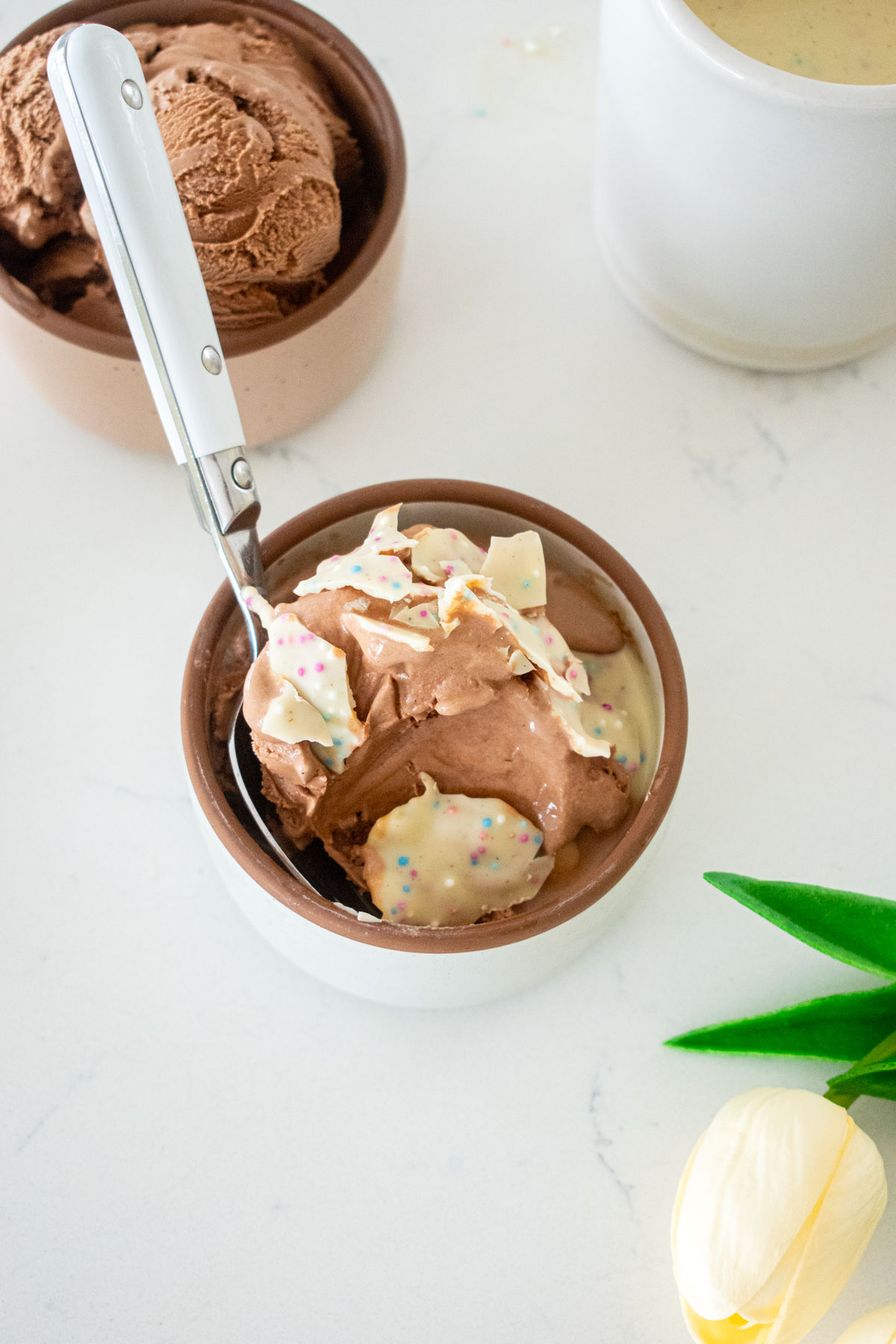 small dish of chocolate ice cream with homemade funfetti magic shell on top crackled with a spoon in the bowl on a marble counter.